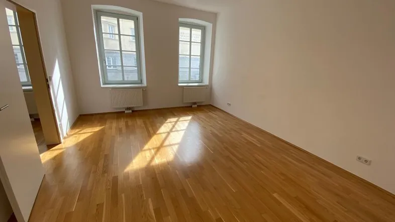 Heller Raum mit Holzboden und zwei Fenstern, durch die Sonnenlicht einfällt.