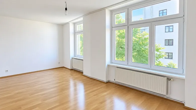 Helles Wohnzimmer mit großen Fenstern, Parkettboden und weiß gestrichenen Wänden.