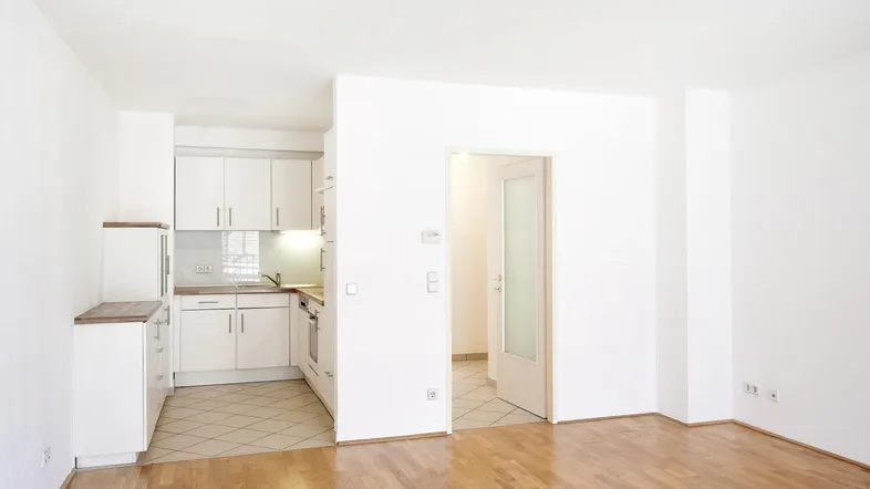 Helles Wohnzimmer mit Parkettboden und offener Küchenzeile, die durch einen Durchgang verbunden ist.
