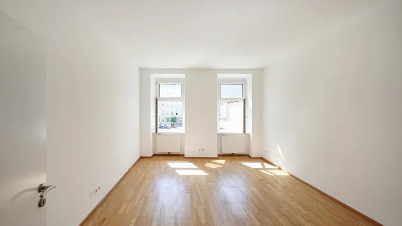 Heller, leerer Raum mit Parkettboden und zwei Fenstern, die viel Tageslicht hereinlassen.