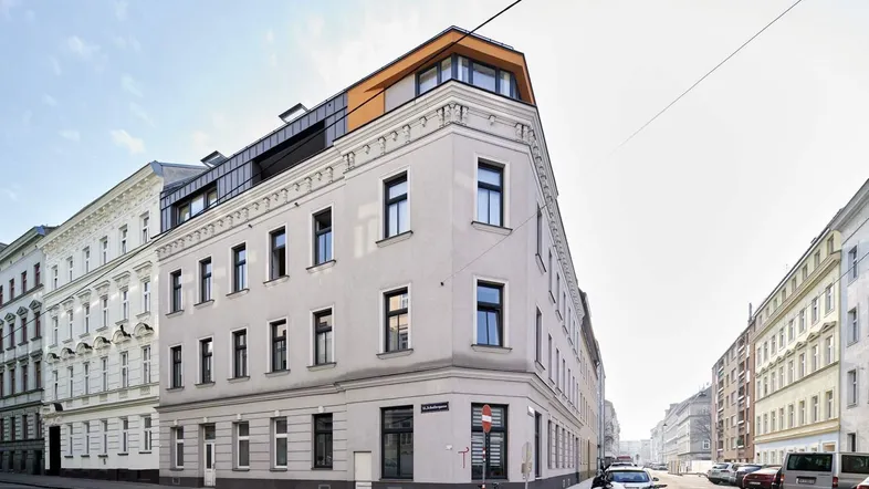 Eckgebäude mit klassischer Fassade und modernem Dachausbau in städtischer Umgebung.