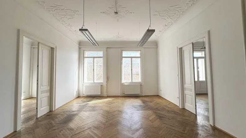 Geräumiges Zimmer mit Fischgrätparkett, hohen Decken und Stuckverzierungen, durchflutet von natürlichem Licht.