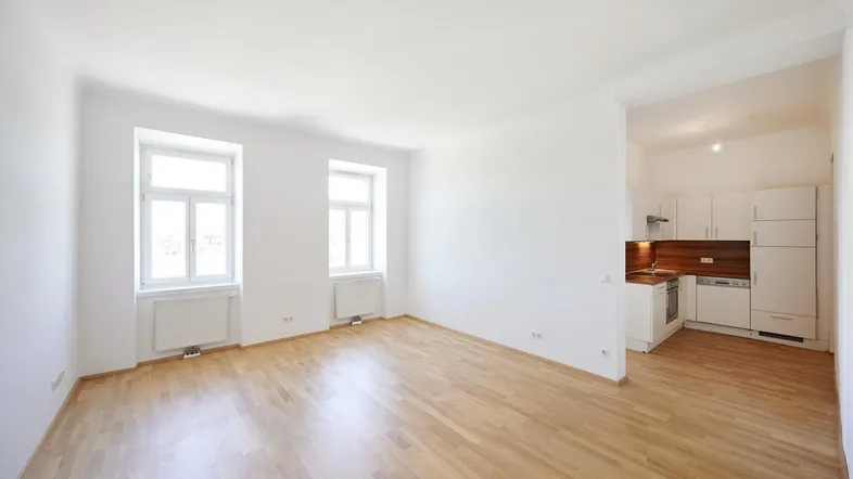 Helles, leeres Zimmer mit Parkettboden und zwei Fenstern, offen zur Küche.