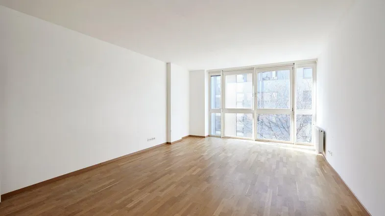 Helles, leeres Zimmer mit Parkettboden und großen Fenstern, die viel Tageslicht hereinlassen.