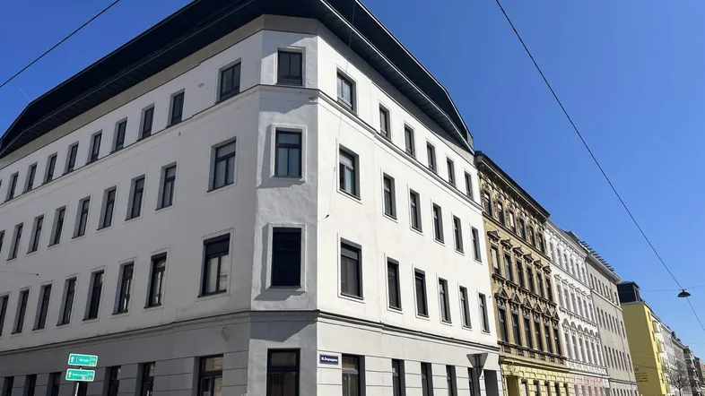 Eckgebäude mit weißer Fassade und dunklen Fenstern unter klarem blauem Himmel.