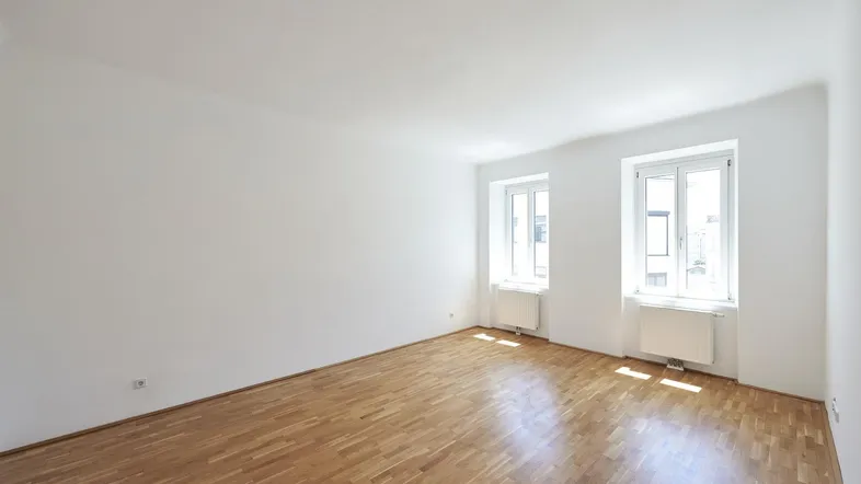 Helles, leeres Zimmer mit Parkettboden und zwei Fenstern, ideal als Wohnbereich nutzbar.