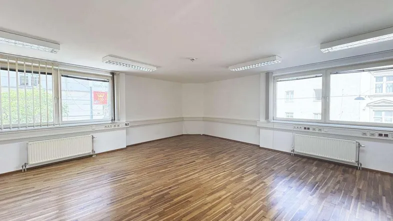 Geräumiges, leeres Büro mit Holzparkettboden, großen Fenstern und effizienter Deckenbeleuchtung.