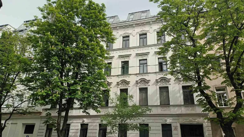 Historische Fassade eines Mehrfamilienhauses mit verzierten Details und grünen Bäumen davor.