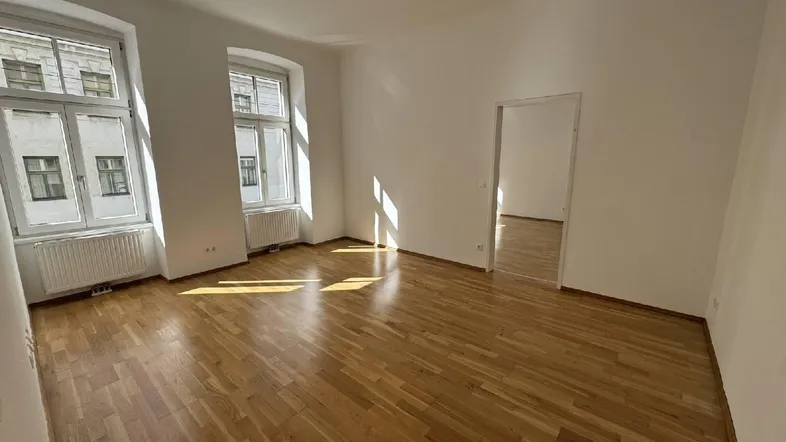 Helles, leeres Zimmer mit Parkettboden und zwei großen Fenstern, das viel Sonnenlicht hereinlässt.