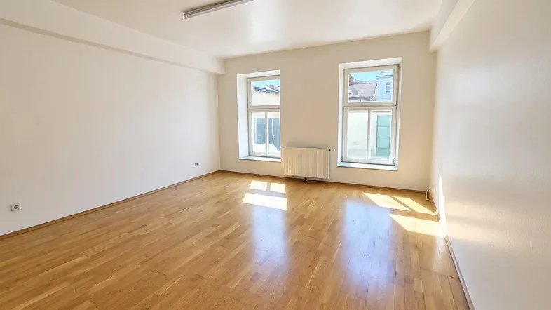 Helles, leeres Zimmer mit Parkettboden und zwei Fenstern, das viel Tageslicht hereinlässt.