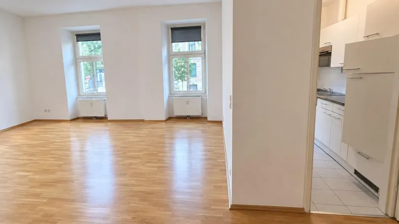 Helles Wohnzimmer mit Parkettboden und zwei großen Fenstern, die viel Tageslicht hereinlassen.