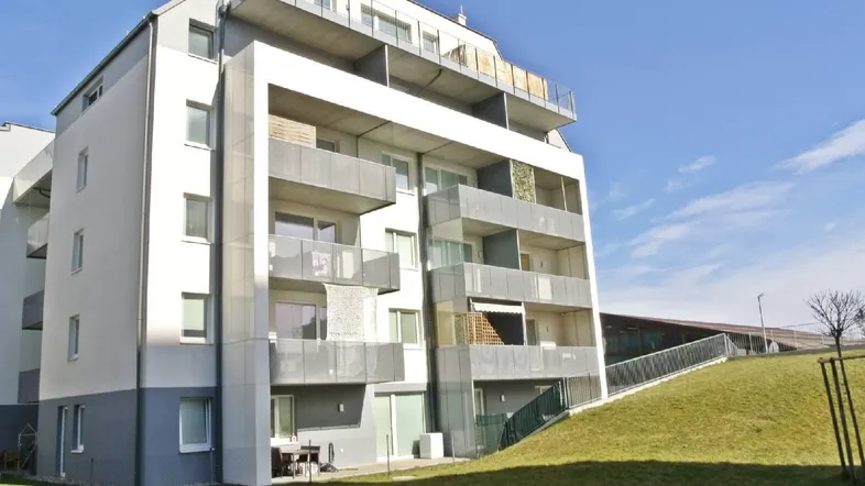 Modernes Mehrfamilienhaus mit Balkonen, Terrassen und Grünflächen unter blauem Himmel.