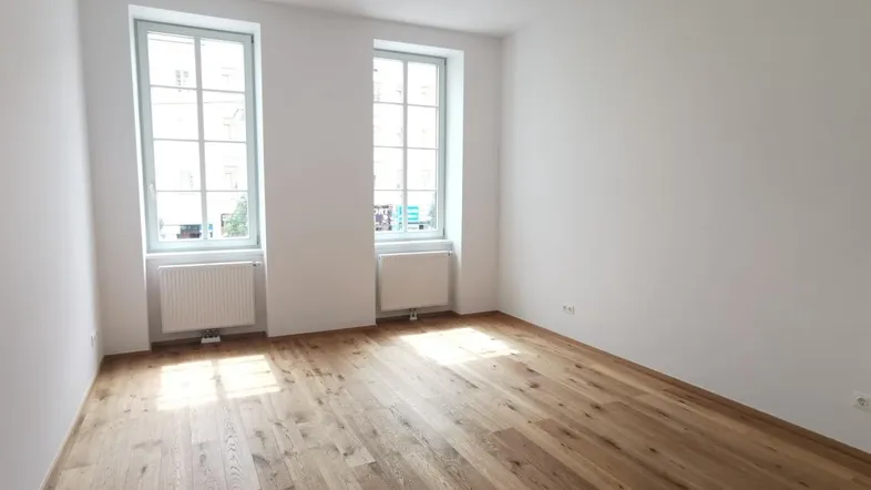 Helles Zimmer mit Holzboden, weißen Wänden und zwei Fenstern, ideal als Wohn- oder Schlafzimmer.