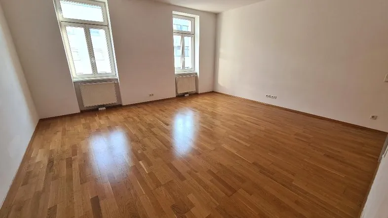 Helles, leeres Zimmer mit glänzendem Parkettboden und zwei großen Fenstern.