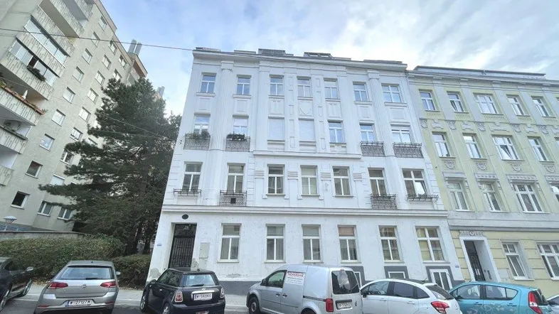 Klassisches Mehrfamilienhaus mit weißer Fassade und kleinen Balkonen an einer belebten Straße.