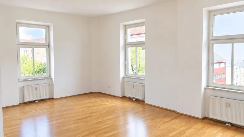 Helles, leeres Zimmer mit Parkettboden und drei Fenstern, bereit zur individuellen Gestaltung.