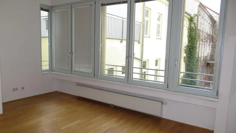 Helles, leeres Zimmer mit Parkettboden und großen Fenstern, die viel Tageslicht hereinlassen.