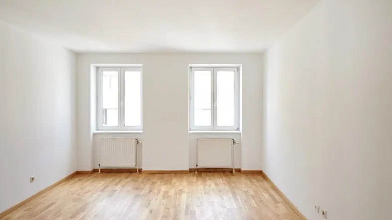 Helles, leeres Zimmer mit Parkettboden und zwei Fenstern, bereit zur individuellen Gestaltung.