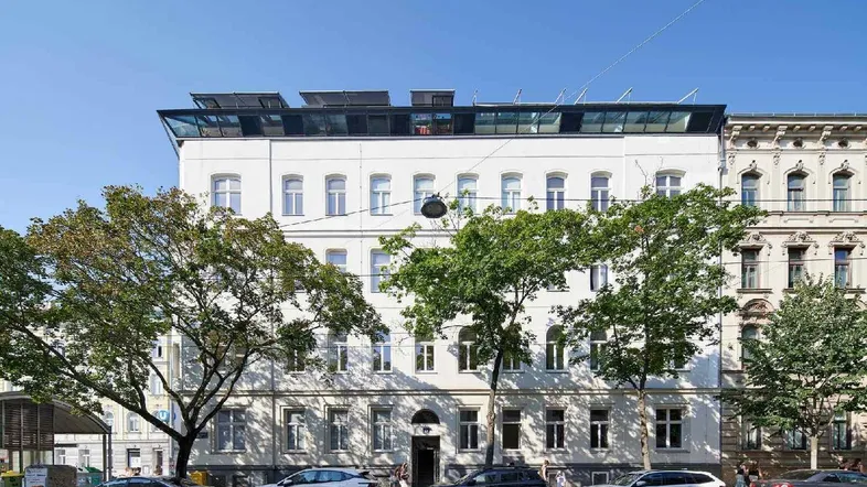Weißes Mehrfamilienhaus mit modernen Dachaufbauten, Photovoltaikanlage und parkenden Autos auf der Straße.