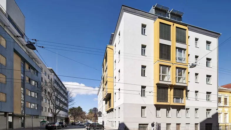Modernes Mehrfamilienhaus an einer Stadtstraße mit gelben Akzenten und blauem Himmel.