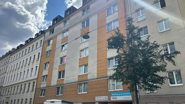 Fassade eines Mehrfamilienhauses mit Mietanzeige und Bäumen an einem sonnigen Tag.