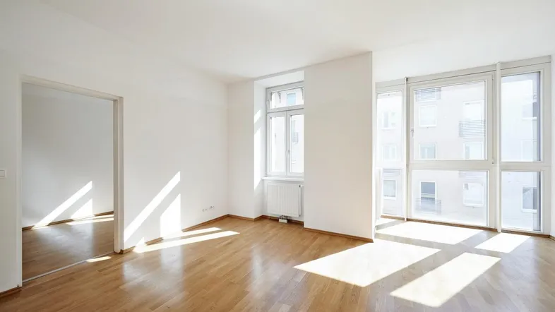 Helles Wohnzimmer mit Parkettboden, weißen Wänden und großen Fenstern, durch die Sonnenlicht fällt.
