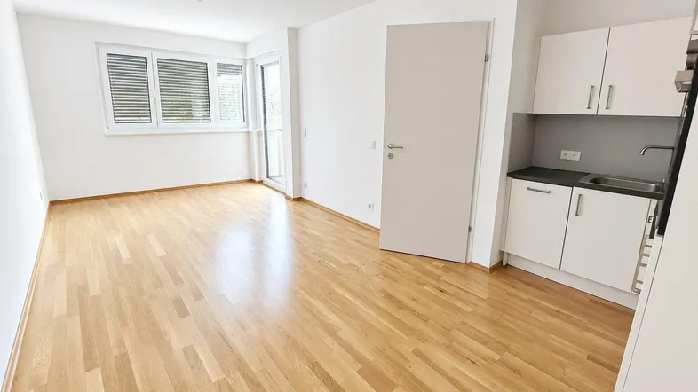 Heller Wohnbereich mit Parkettboden, integrierter Küchenzeile und Zugang zu einem Balkon.