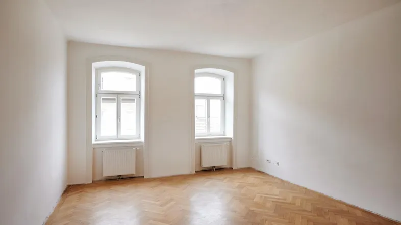 Helles Zimmer mit Fischgrätparkett und zwei Fenstern, bereit zur individuellen Gestaltung.