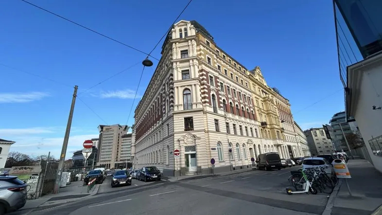 Historisches Gebäude an einer Straßenecke mit klassischer Architektur und geparkten Fahrzeugen unter blauem Himmel.