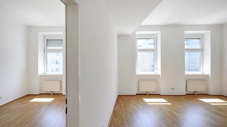 Helles, leeres Zimmer mit Parkettboden und mehreren Fenstern, das viel Tageslicht hereinlässt.