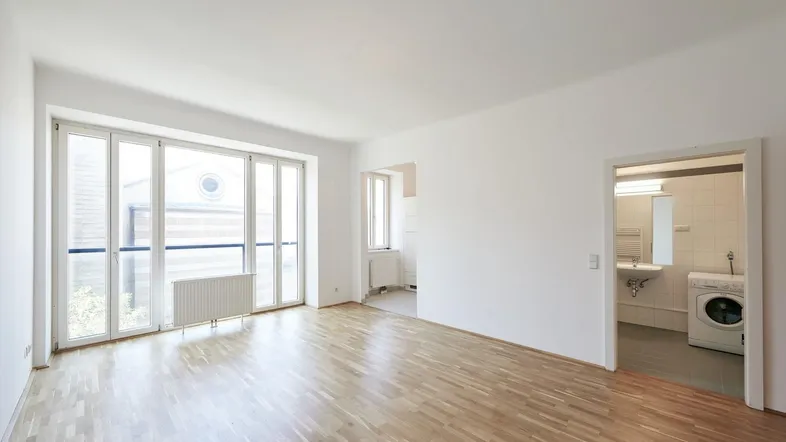 Helles, leeres Zimmer mit Parkettboden und großen Fenstern, das viel Tageslicht hereinlässt.