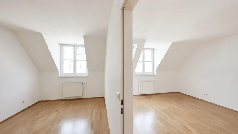 Helles Zimmer im Dachgeschoss mit Dachschrägen, Holzparkettboden und Fenstern.