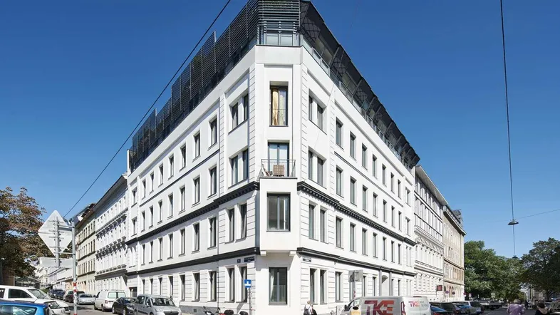 Modernes Eckgebäude mit weißer Fassade und vielen Fenstern, umgeben von städtischer Umgebung.