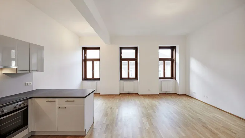 Helles Wohnzimmer mit integrierter Küchenzeile, Parkettboden und drei großen Fenstern.