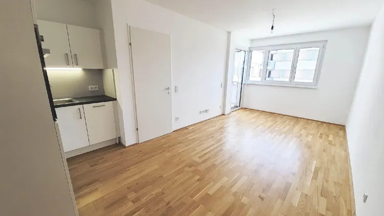 Helles Wohnzimmer mit integrierter Küchenzeile, Parkettboden und Zugang zu einem Balkon.