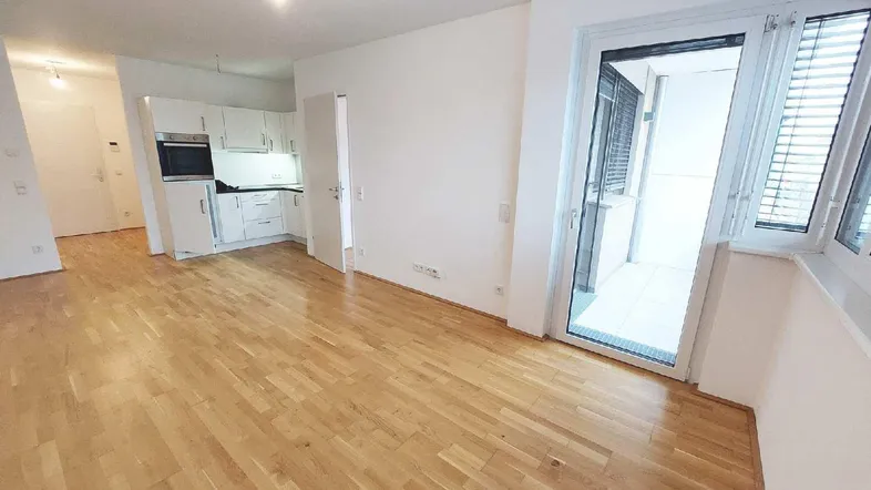 Heller Wohnbereich mit Parkettboden, integrierter Küchenzeile und Zugang zu einem Balkon.