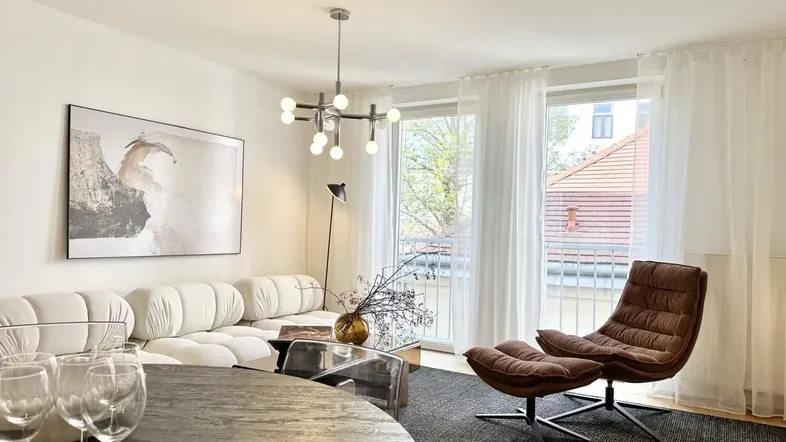 Helles Wohnzimmer mit modernem Sofa, bequemen Sessel und Zugang zu einem kleinen Balkon.