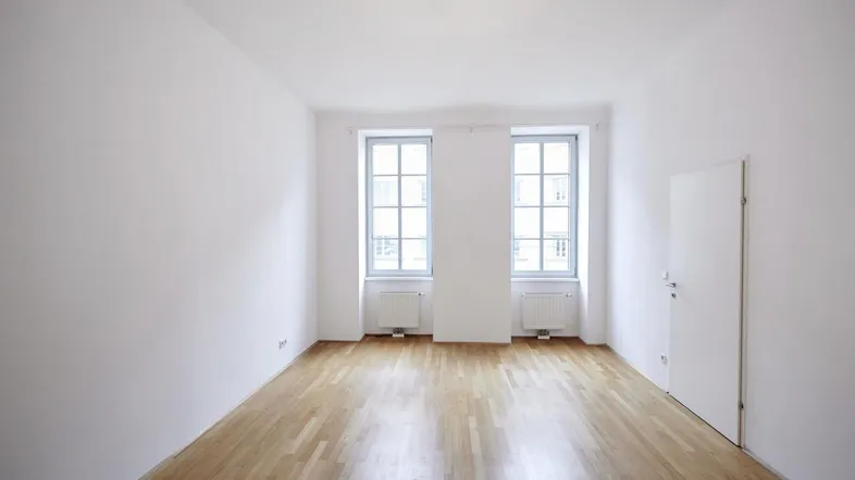Helles, leeres Zimmer mit Parkettboden und zwei Fenstern, bereit zur individuellen Gestaltung.