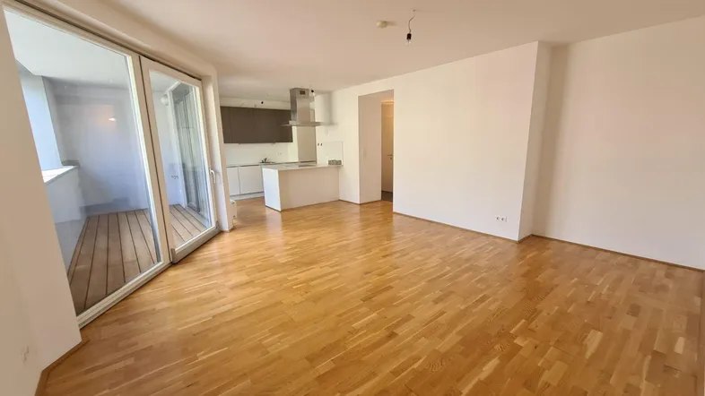 Heller offener Wohnbereich mit Parkettboden, integrierter Küche und Zugang zu einem Balkon.