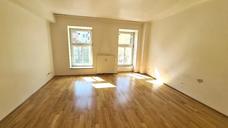 Helles Zimmer mit Parkettboden, weißen Wänden und zwei Fenstern, das viel Tageslicht hereinlässt.