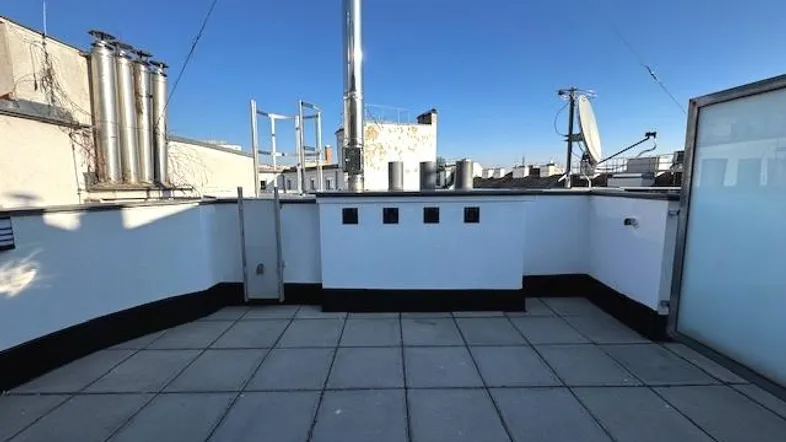Geräumige Dachterrasse mit Fliesenboden und weitem Blick über die Dächer der Stadt unter blauem Himmel.