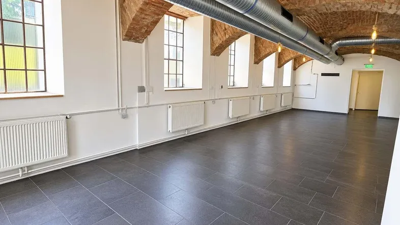 Großer, offener Raum mit Gewölbedecke, sichtbaren Lüftungsrohren und großen Fenstern.
