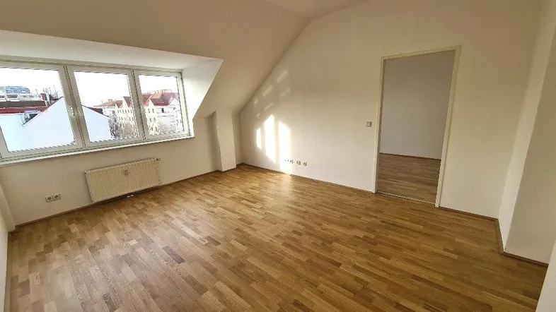 Helles Zimmer im Dachgeschoss mit Holzparkettboden, schrägen Wänden und einem großen Fenster.
