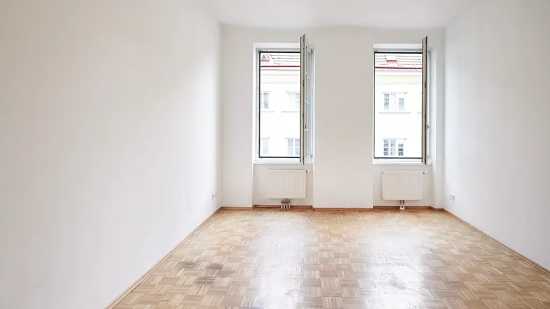 Helles Zimmer mit Parkettboden und zwei Fenstern, die viel Tageslicht hereinlassen.
