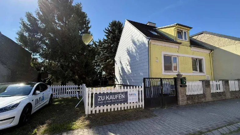 Gelbes Einfamilienhaus mit weißem Zaun und einem "Zu Kaufen" Schild an der Straße.
