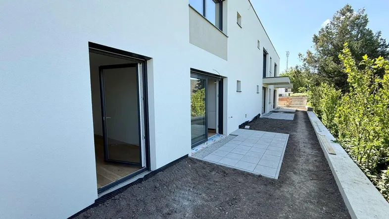 Moderne Reihenhausfassade mit weißem Putz und einer neu angelegten Terrasse, umgeben von viel Grün.