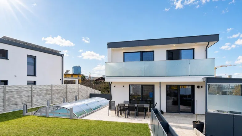 Modernes Einfamilienhaus mit großem Garten, Terrasse und überdachtem Pool unter strahlend blauem Himmel.