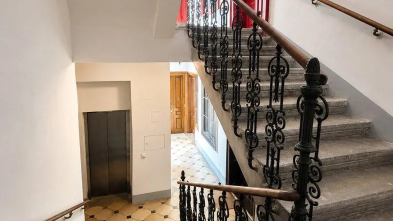 Historisches Treppenhaus mit schmiedeeisernem Geländer und Zugang zu einem modernen Aufzug.