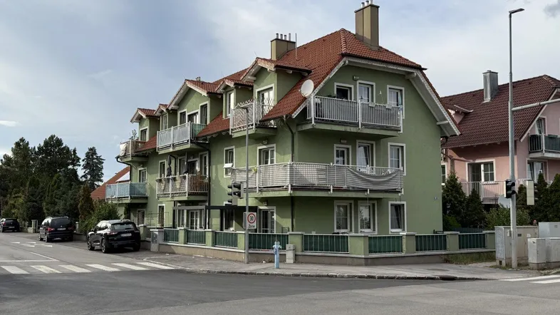 Mehrfamilienhaus mit grüner Fassade, roten Ziegeldächern und mehreren Balkonen an einer Straßenecke.