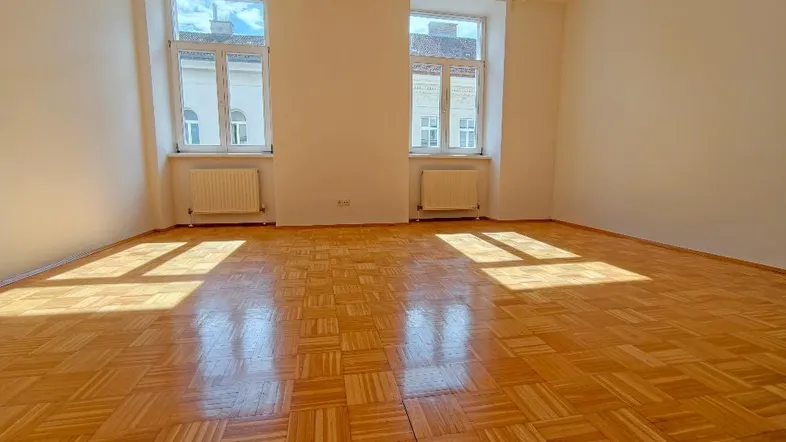 Helles Zimmer mit Parkettboden, weißen Wänden und zwei Fenstern, durch die Sonnenlicht einfällt.